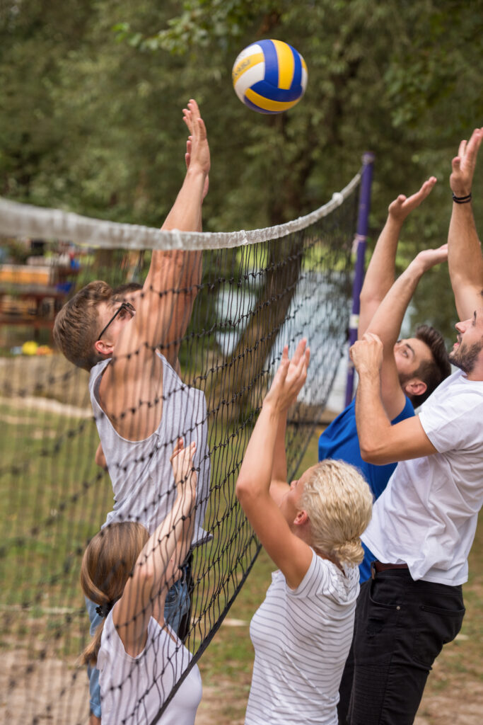 Gruppe junger Freunde spielt Beach-Volleyball im Freien am Netz; gelber/blauer Volleyball in der Luft, umgeben von Bäumen im Fichtelgebirge, Oberfranken