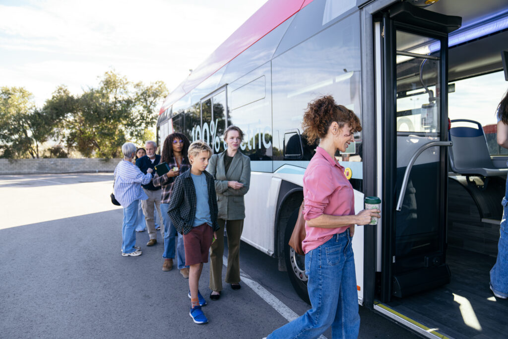 Gruppe von Reisenden wartet vor einem elektrischen Bus, mehrere Personen stehen Schlange, Bus zeigt 100% electric-Aufschrift, Outdoor-Szene