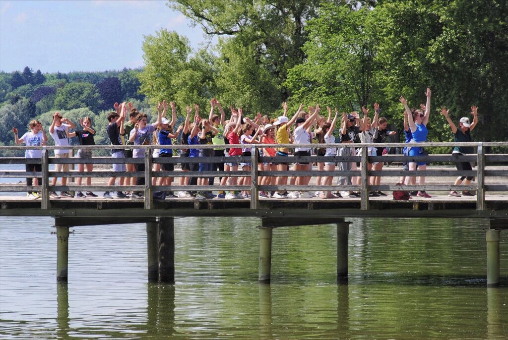 Schülergruppe mit erhobenen Armen auf einer Holzbrücke über einem See bei einer abwechslungsreichen Klassenreise im Grünen