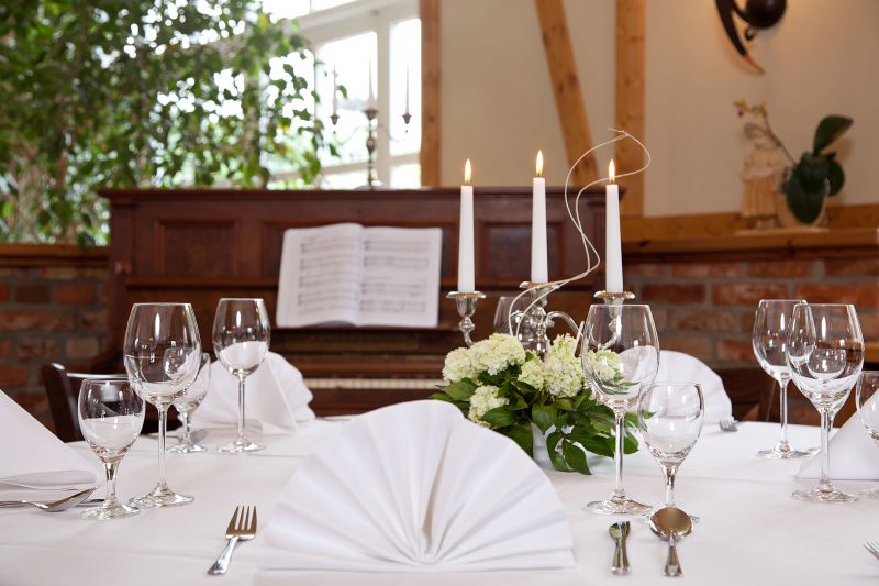 Festlich gedeckter Restauranttisch im Landhof Liepe mit weißen Servietten, Blumen, Kerzen und Gläsern vor einem Klavier.