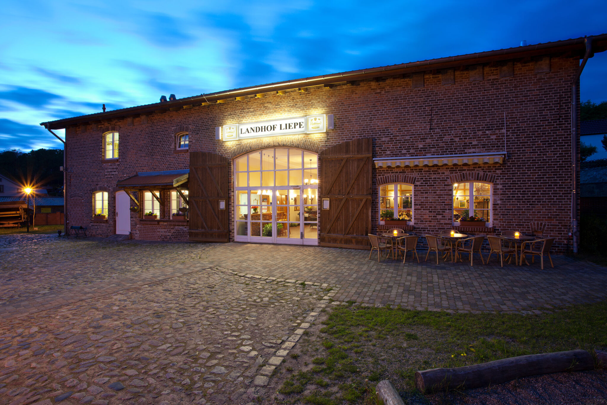 Das Restaurant Landhof Liepe zeigt sich abends als rustikantes Backsteingebäude mit beleuchtetem Eingang und Außenterrasse.
