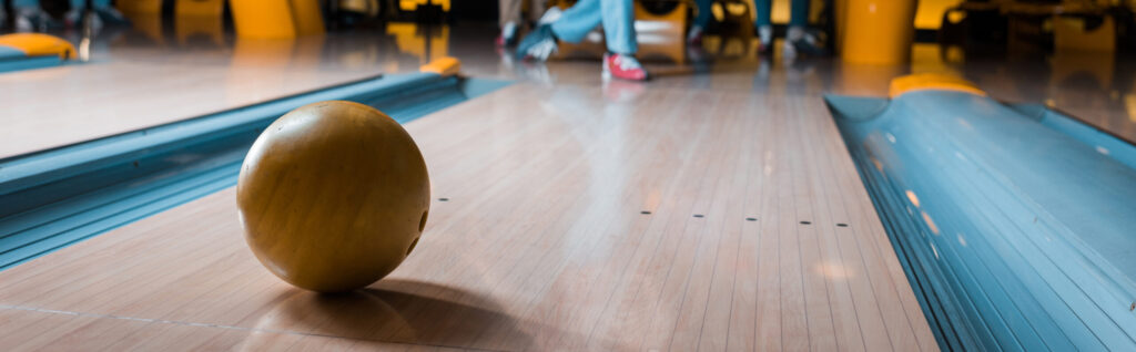 Auf der hölzernen Kegelbahn liegt eine braune Bowlingkugel; am anderen Ende steht eine Person in blauer Hose und pinken Schuhen.